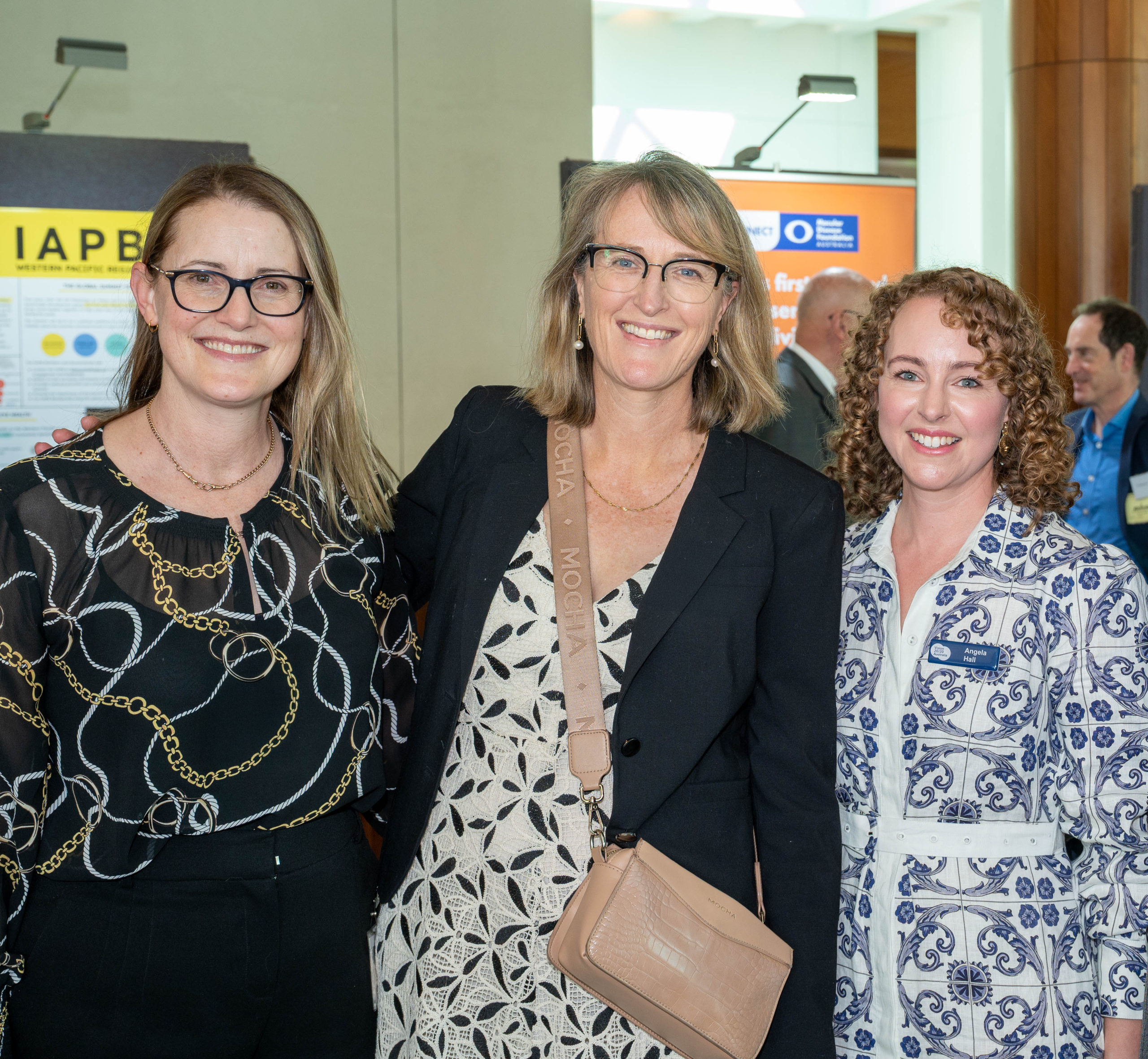 Three women stand together, smiling at a conference. They wear professional attire with vibrant patterns.