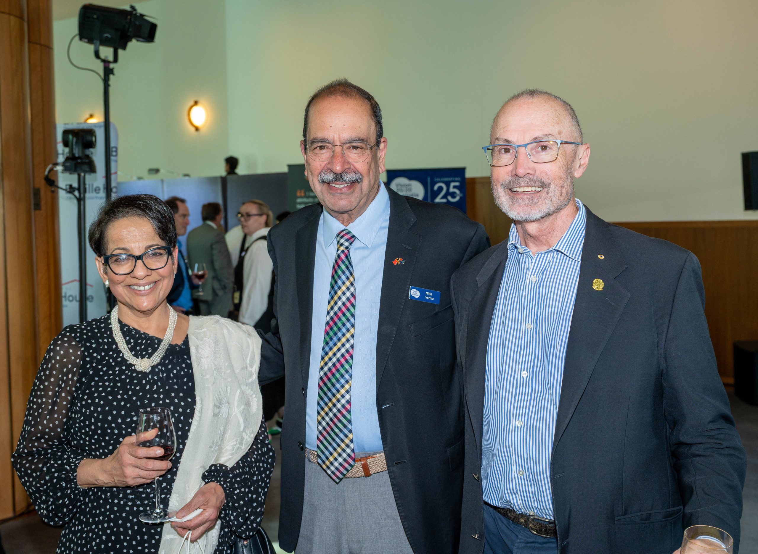 Three people are smiling in a formal indoor setting. One woman holds a wine glass. They appear relaxed and joyful, dressed in smart attire.